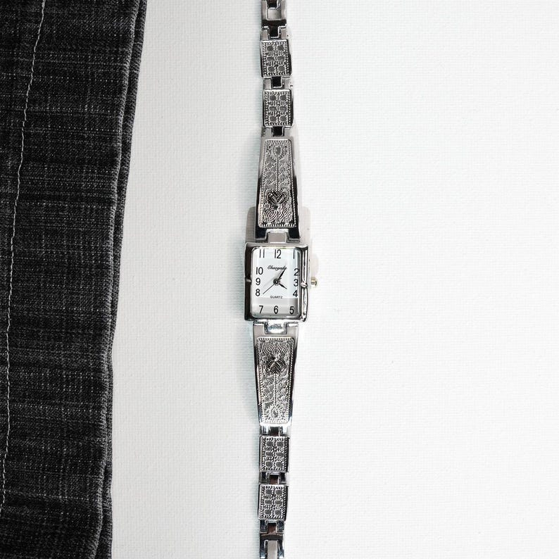 May include: A silver-toned rectangular watch with a matching bracelet. The watch face displays the time with black numerals and hands. The bracelet and watch casing feature decorative patterns.