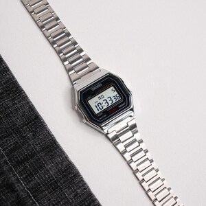 Puede incluir: Reloj digital plateado con esfera rectangular y correa a juego. La esfera muestra la hora, 10:33:36. El reloj es resistente al agua hasta 30m. La marca "SKEI" es visible.