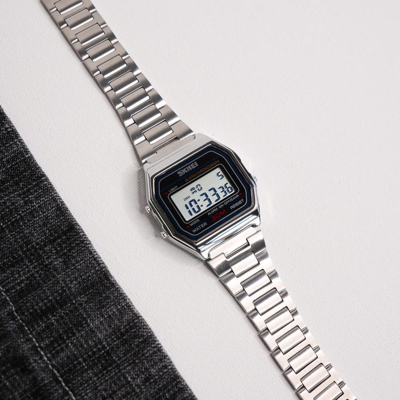 Puede incluir: Reloj digital plateado con esfera rectangular y correa a juego. La esfera muestra la hora, 10:33:36. El reloj es resistente al agua hasta 30m. La marca "SKEI" es visible.