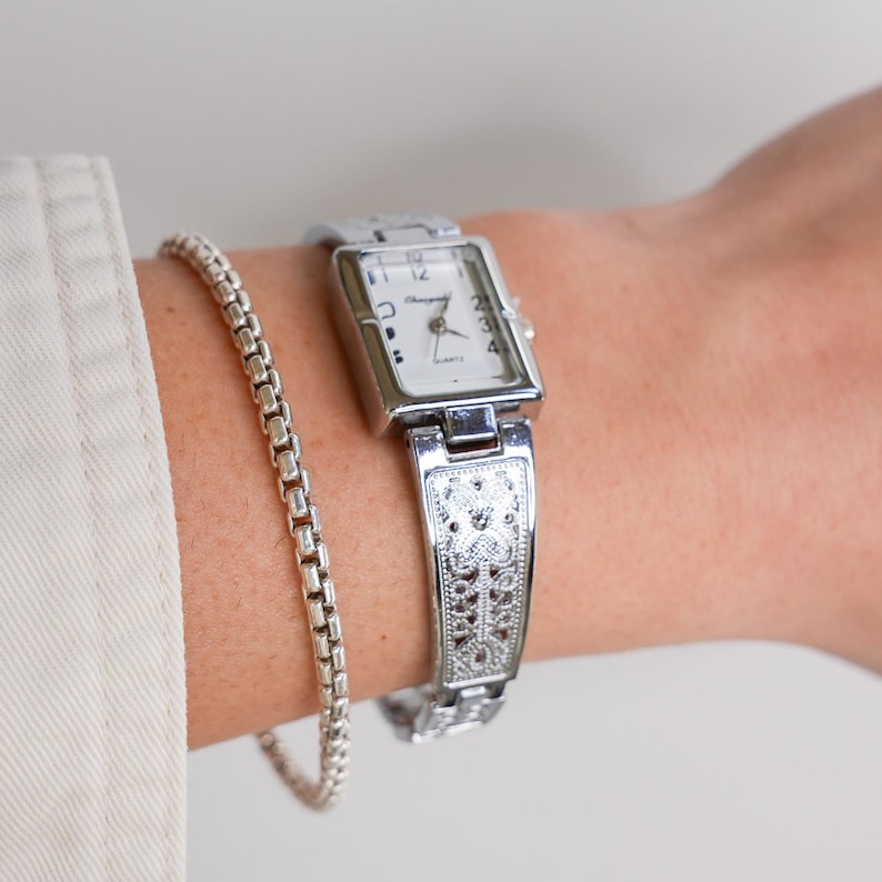 May include: A silver-toned rectangular watch with a white face and black numerals is worn on a wrist. The watch has an ornate band. A silver chain bracelet is also on the wrist. The watch face reads "Changmei Quartz".