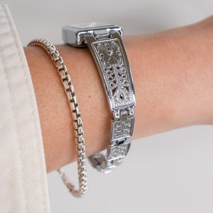 May include: A silver-toned watch with a rectangular face and an ornate, openwork band is shown on a wrist. A silver chain bracelet is also visible. The watch band features a detailed, symmetrical design. The watch and bracelet are both reflective.
