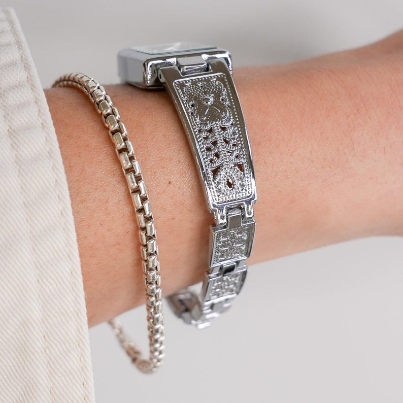 May include: A silver-toned watch with a rectangular face and an ornate, openwork band is shown on a wrist. A silver chain bracelet is also visible. The watch band features a detailed, symmetrical design. The watch and bracelet are both reflective.
