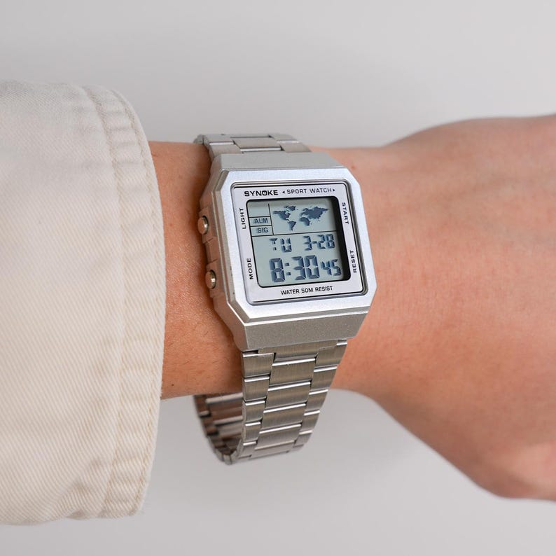 May include: A silver digital watch with a rectangular face and a matching metal band. The watch displays the time, date, and a world map icon. The watch is labeled "SYNOKE SPORT WATCH" and "WATER 50M RESIST".