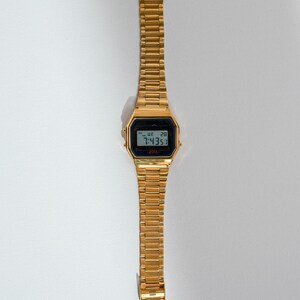 May include: A gold-toned digital wristwatch with a rectangular black display showing the time and date. The watch has a matching gold-colored metal band. The watch is displayed on a white surface.