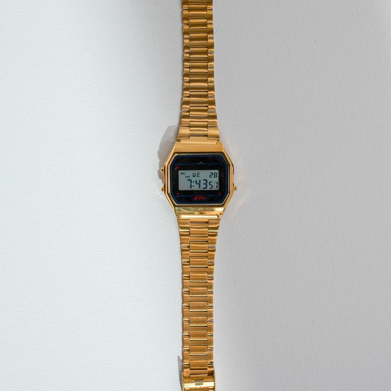 May include: A gold-toned digital wristwatch with a rectangular black display showing the time and date. The watch has a matching gold-colored metal band. The watch is displayed on a white surface.