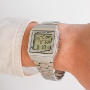 May include: A silver-toned digital sport watch with a rectangular face and matching metal band. The watch displays the time, date, and world map icon. The watch face has the text "SYNOKE SPORT WATCH" and "WATER 50M RESIST".