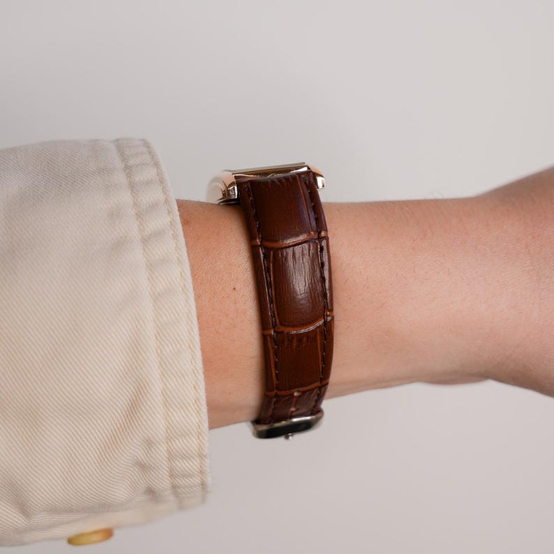 May include: A brown leather watch band with a rectangular watch face. The watch band has a crocodile skin texture. The watch face is gold and silver. The watch is worn on a wrist.