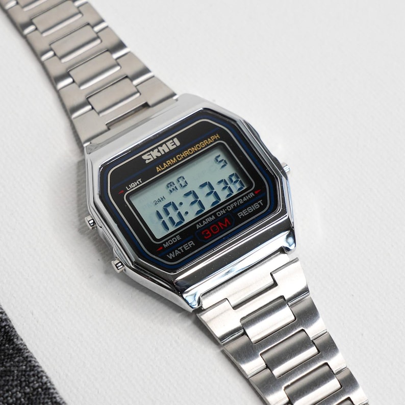 Puede incluir: Reloj digital plateado con esfera rectangular y correa met&aacute;lica a juego. La pantalla muestra la hora 10:33:39. La marca "SKMEI" es visible. Funciones: alarma y resistencia al agua hasta 30 metros.