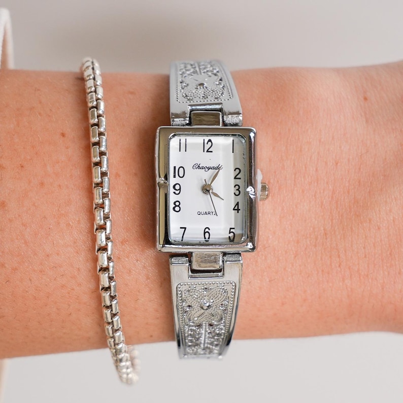 May include: A silver-toned rectangular watch with a white face and black numerals. The watch band and face frame feature an ornate, engraved design. A silver bracelet is visible next to the watch.