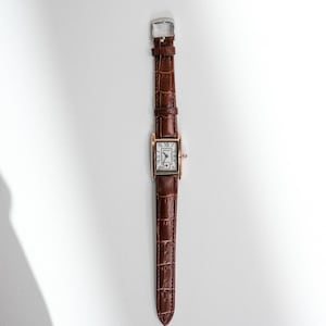 May include: A rectangular wristwatch with a brown leather band and a gold-colored case. The watch face is white with black Roman numerals. The band has a crocodile texture and a silver buckle.