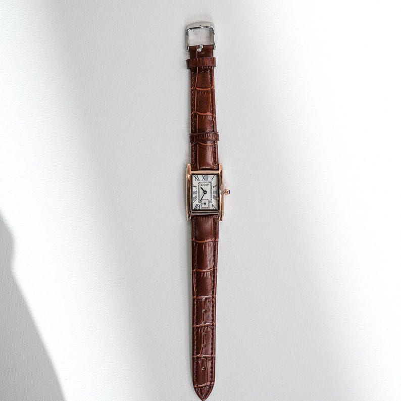 May include: A rectangular wristwatch with a brown leather band and a gold-colored case. The watch face is white with black Roman numerals. The band has a crocodile texture and a silver buckle.