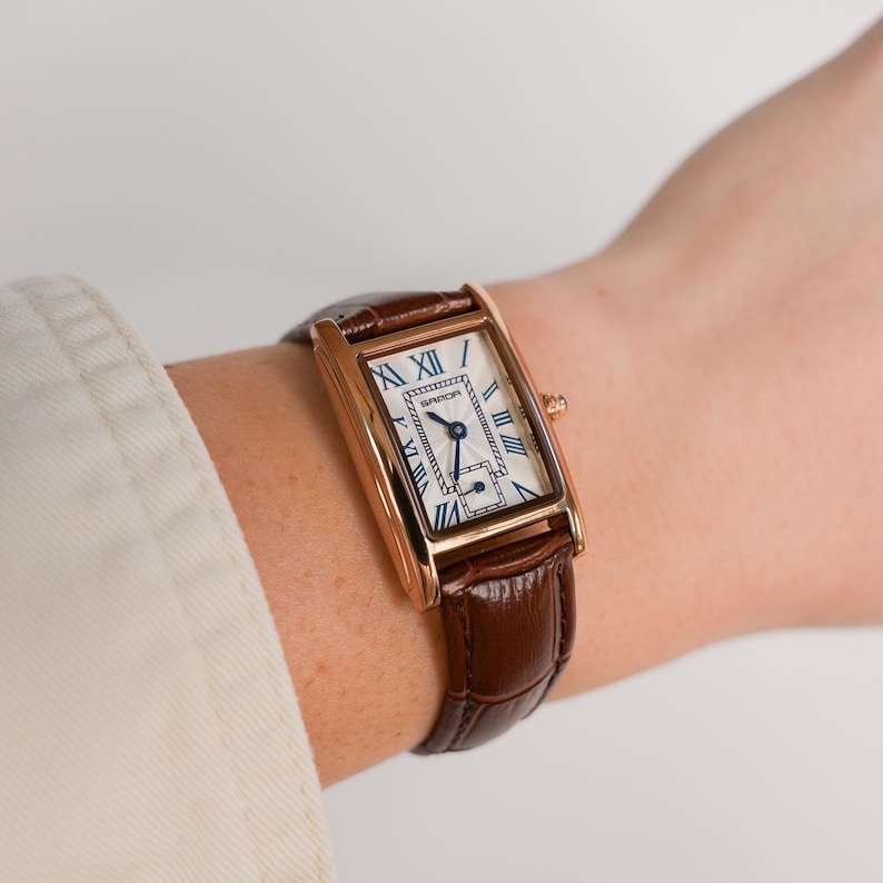 May include: A rectangular gold-tone wristwatch with a brown leather band. The watch face is white with blue Roman numerals and hands. The band is a dark brown, textured leather. The watch is on a wrist.