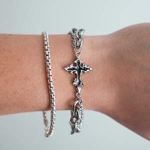 May include: Two silver bracelets are displayed. One is a simple chain, and the other features a decorative cross pendant with black enamel detailing. The cross is suspended from a chain with ornate links. The bracelets are shown on a person's arm.