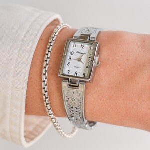 May include: A silver-toned quartz watch with a rectangular face and a matching bracelet-style band. The watch face is white with black numerals and hands. The band and watch case feature an ornate, engraved design. A silver chain bracelet is also visible.