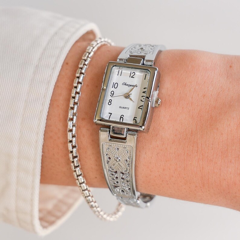 May include: A silver-toned quartz watch with a rectangular face and a matching bracelet-style band. The watch face is white with black numerals and hands. The band and watch case feature an ornate, engraved design. A silver chain bracelet is also visible.