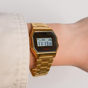 May include: A gold-toned digital watch with a rectangular face and matching band is displayed. The watch face shows the time as 11:13:35. The watch is worn on a wrist, with a white shirt sleeve visible in the background.