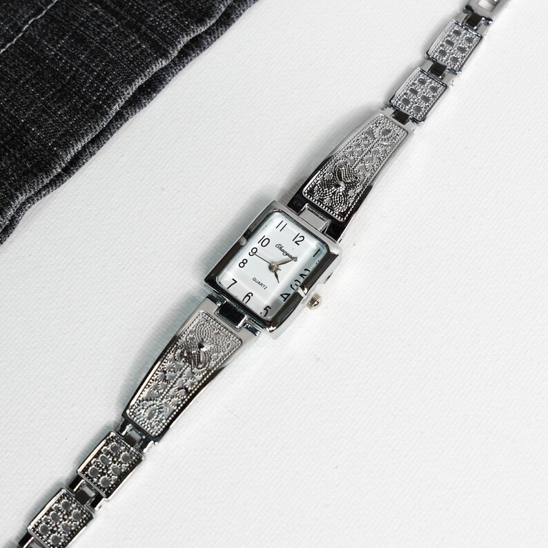 May include: A silver-toned quartz watch with an ornate, engraved band. The rectangular watch face has a white background with black numerals and hands. The watch band features detailed floral and geometric patterns.