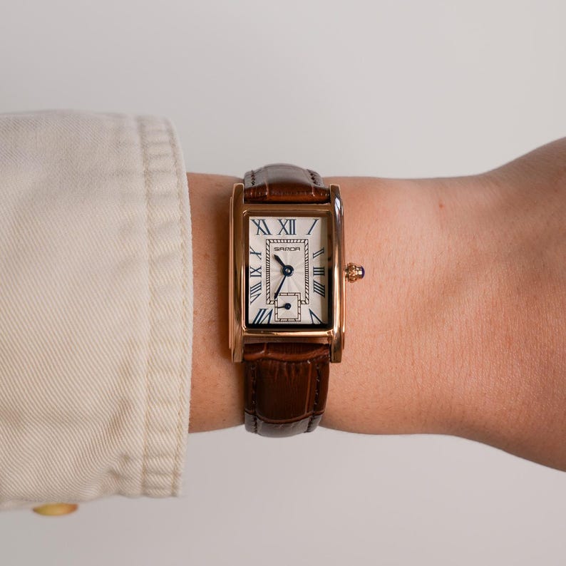 May include: A rectangular gold-tone wristwatch with a brown leather band. The watch face is white with black Roman numerals and hands. The watch is secured on a wrist.