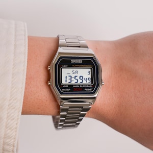 Puede incluir: Un reloj digital SKMEI plateado con una esfera rectangular y una correa de metal a juego. El reloj muestra la hora, el d&iacute;a y la fecha en una pantalla en blanco y negro. El reloj es resistente al agua hasta 30 metros.