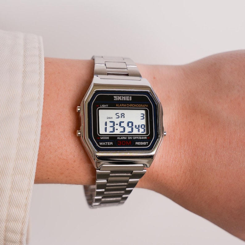 Puede incluir: Un reloj digital SKMEI plateado con una esfera rectangular y una correa de metal a juego. El reloj muestra la hora, el d&iacute;a y la fecha en una pantalla en blanco y negro. El reloj es resistente al agua hasta 30 metros.