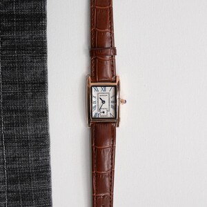 May include: A rectangular gold-toned wristwatch with a brown leather band. The watch face is white with blue Roman numerals and hands. The band has a crocodile texture.