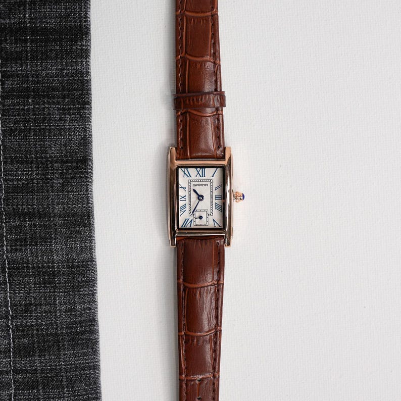 May include: A rectangular gold-toned wristwatch with a brown leather band. The watch face is white with blue Roman numerals and hands. The band has a crocodile texture.