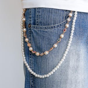 May include: A decorative belt chain with two strands. One strand is a string of white pearls, and the other is a gold chain with pearls and colorful gemstones. The chain is attached to a pair of blue denim jeans.