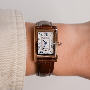 May include: A rectangular gold-toned wristwatch with a white face and blue hands and Roman numerals. The watch has a brown leather band. The watch face has the brand name "SARDA" printed on it.