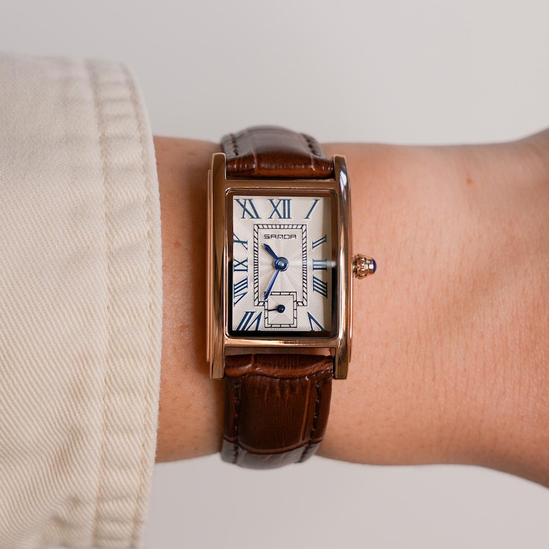 May include: A rectangular gold-toned wristwatch with a white face and blue hands and Roman numerals. The watch has a brown leather band. The watch face has the brand name "SARDA" printed on it.