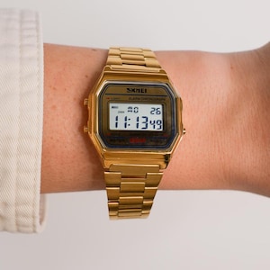 May include: Gold-toned digital watch with a rectangular face and matching link band. The watch displays the time as 11:13:49. The watch face has the brand name "SKMEI" and other text. The watch is on a wrist.