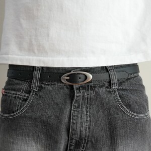 May include: Black belt with a silver oval buckle, worn with gray denim jeans and a white t-shirt. The belt is made of a smooth, dark material and has a simple, classic design. The jeans have white stitching and a classic five-pocket design.