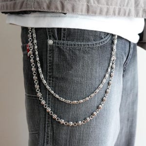 May include: Silver-toned metal wallet chain with two strands, one with a series of small, decorative crosses and the other with a series of oval links. The chain is attached to a pair of gray denim jeans.