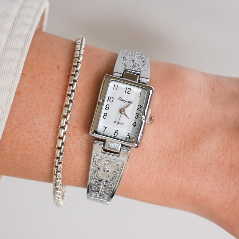 May include: Silver-toned rectangular quartz watch with a white face and black numerals. The watch has decorative detailing on the band and a silver bracelet. The watch face reads "Chaoyade Quartz".