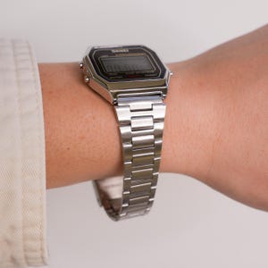 Puede incluir: Se muestra un reloj digital plateado con esfera negra y correa plateada a juego. La esfera del reloj muestra la hora. El reloj est&aacute; en una mu&ntilde;eca y la correa est&aacute; hecha de eslabones rectangulares.