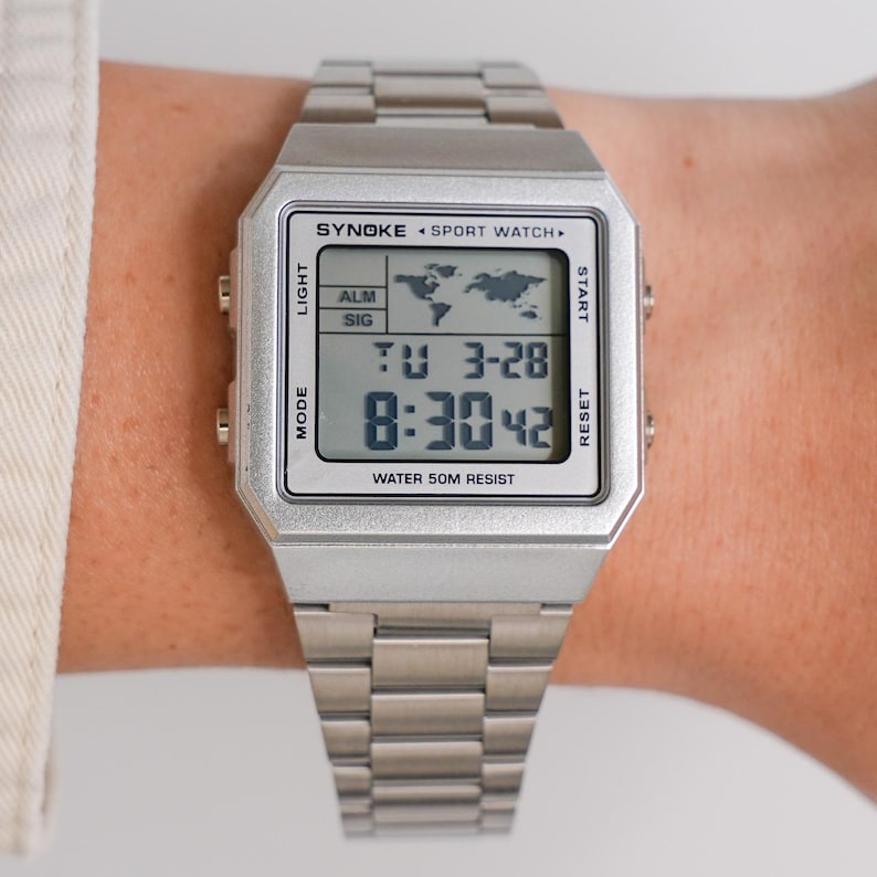 May include: A silver-toned digital sport watch with a rectangular face and a matching metal band. The watch face displays the time, date, and a world map graphic. The text "SYNOKE SPORT WATCH" is visible, along with "WATER 50M RESIST".