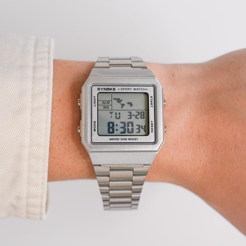 May include: A silver digital sport watch with a rectangular face and a matching metal band. The watch face displays the time, date, and other functions. The watch has the text "SYNOKE SPORT WATCH" and "WATER 50M RESIST".