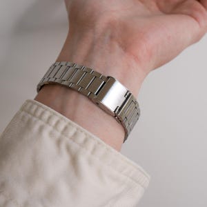 May include: A silver-toned metal watch band is clasped on a wrist. The watch band is made of rectangular links and has a secure clasp. The person is wearing a light-colored long-sleeved shirt.