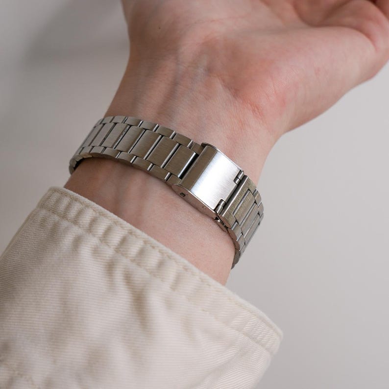 May include: A silver-toned metal watch band is clasped on a wrist. The watch band is made of rectangular links and has a secure clasp. The person is wearing a light-colored long-sleeved shirt.