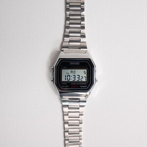 Puede incluir: Un reloj digital plateado con una esfera negra rectangular que muestra la hora 10:33:27. El reloj tiene una correa de acero inoxidable y la marca "SKMEI" es visible. El reloj es resistente al agua hasta 30 metros.