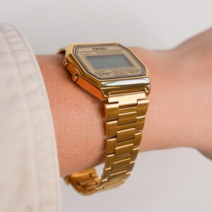 May include: A gold-toned digital watch with a rectangular face and matching metal band. The watch displays the time on a digital screen and features the brand name "SKEI". The watch is water-resistant up to 30 meters.