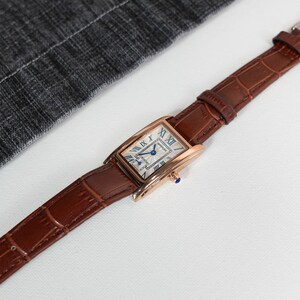 May include: A rectangular rose gold wristwatch with a brown leather band. The watch face is white with blue Roman numerals and hands. The band has a crocodile texture.