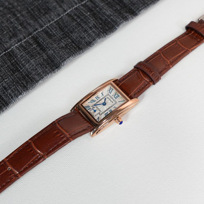 May include: A rectangular rose gold wristwatch with a brown leather band. The watch face is white with blue Roman numerals and hands. The band has a crocodile texture.