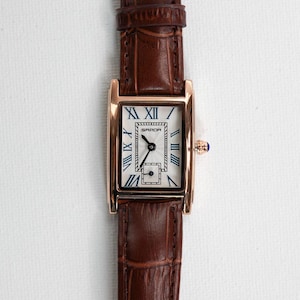 May include: A rectangular wristwatch with a brown leather band and a gold-colored case. The watch face is white with black Roman numerals and blue hands. The watch has a small, square subdial and a blue crown.