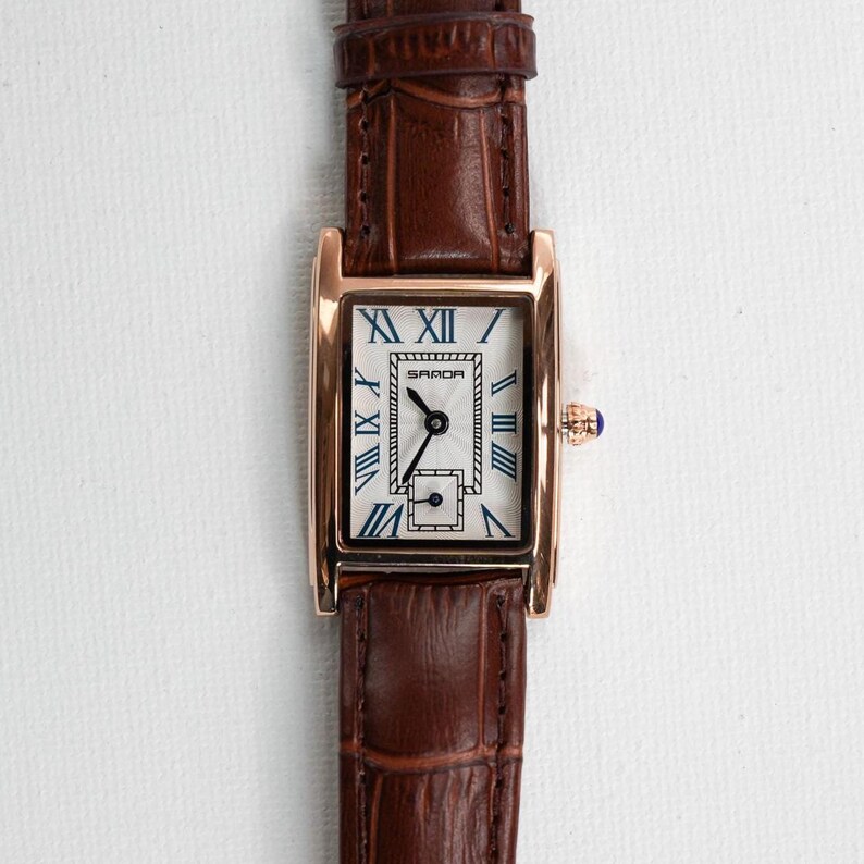 May include: A rectangular wristwatch with a brown leather band and a gold-colored case. The watch face is white with black Roman numerals and blue hands. The watch has a small, square subdial and a blue crown.