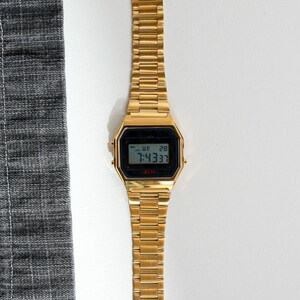 May include: A gold-toned digital watch with a rectangular face and matching band. The watch displays the time, day, and date on a black screen. The watch is set to 7:43:37. The watch is on a white surface next to a gray fabric.