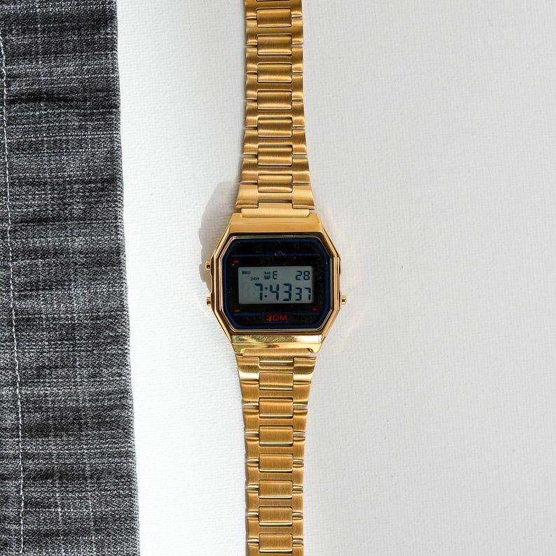 May include: A gold-toned digital watch with a rectangular face and matching band. The watch displays the time, day, and date on a black screen. The watch is set to 7:43:37. The watch is on a white surface next to a gray fabric.