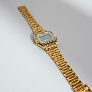May include: A gold-toned digital watch with a rectangular face and a matching link band. The watch face displays the time and has the brand name "SKEI" printed on it. The watch is angled, showing its details.