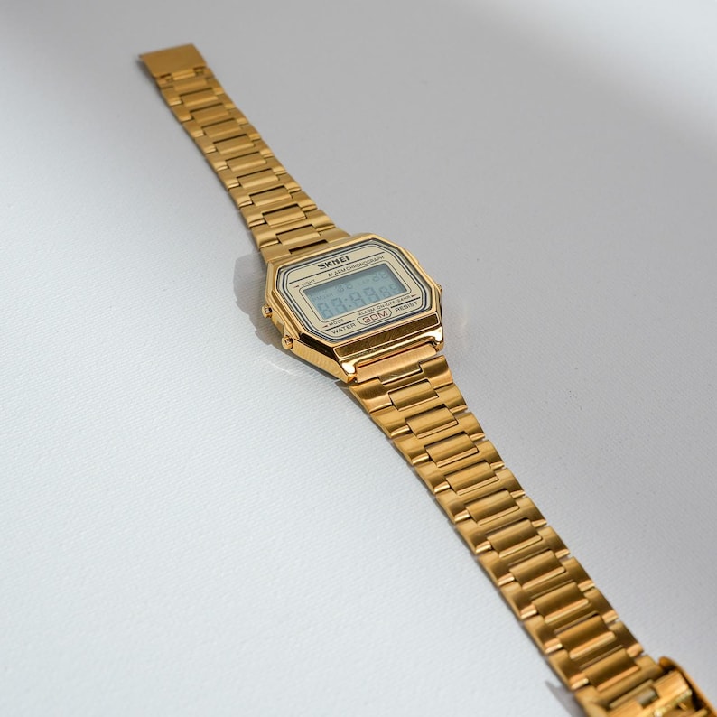 May include: A gold-toned digital watch with a rectangular face and a matching link band. The watch face displays the time and has the brand name "SKEI" printed on it. The watch is angled, showing its details.