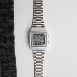 May include: A silver-toned digital watch with a rectangular face and a matching metal band. The watch face displays the time and date, with the text "SYNOKE SPORT WATCH" visible. The watch is water resistant.