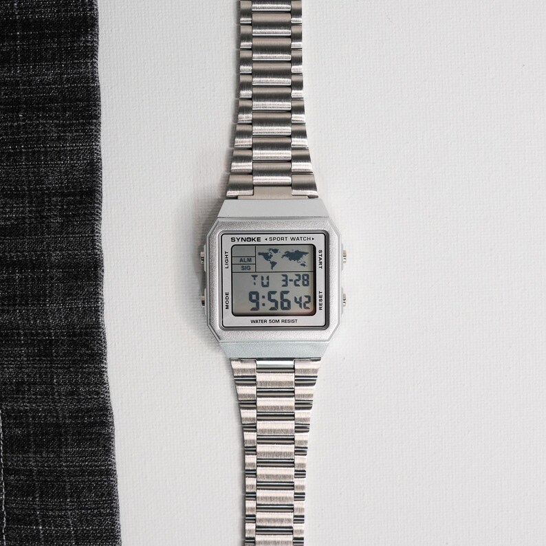 May include: A silver-toned digital watch with a rectangular face and a matching metal band. The watch face displays the time and date, with the text "SYNOKE SPORT WATCH" visible. The watch is water resistant.
